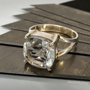 May include: A silver ring featuring a large, square, clear gemstone. The ring has a thick band and decorative prongs securing the stone. It rests on a dark, textured surface. The text "JewelsByEkta" is visible.