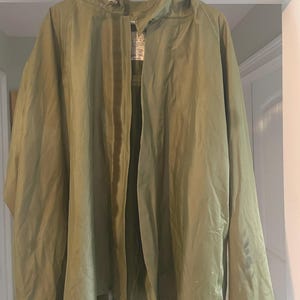 May include: An olive green hooded jacket hanging on a white hanger. The jacket is open, revealing a tag with text. The jacket has a button closure and appears to be made of a lightweight material. The jacket is hanging in front of a white door.