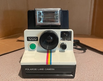 Può includere: Una fotocamera Polaroid Land Camera modello 1000 vintage, con corpo bianco e parte superiore nera. Presenta una striscia arcobaleno e la scritta "POLAROID LAND CAMERA". È visibile un pulsante verde. Una classica fotocamera istantanea.
