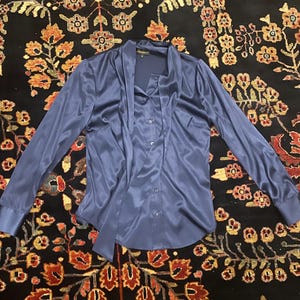 May include: A long-sleeved, navy blue satin blouse with a draped neckline and button-down front. The blouse features a tie detail at the neck and buttoned cuffs. The garment is displayed on a patterned rug.