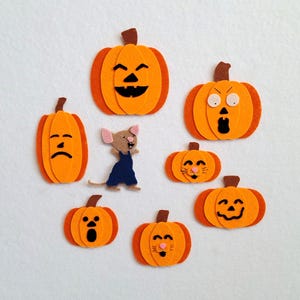 Op de afbeelding: Een verzameling oranje vilten pompoenen met verschillende gezichtsuitdrukkingen, waaronder blij, verdrietig en verrast. Een kleine muis met een blauwe overall is ook inbegrepen. De pompoenen zijn een feestelijke decoratie voor Halloween.