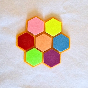 May include: A colorful felt coaster set in a honeycomb design. The coaster features seven hexagonal pieces in various bright colors: pink, yellow, blue, orange, green, purple, and red, set within a yellow border. The background is white.