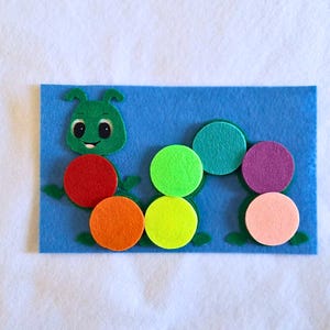 May include: A colorful felt caterpillar toy on a blue felt background. The caterpillar has a green head with a smiling face and a series of colorful circular body segments in red, orange, green, yellow, teal, purple, and pink.