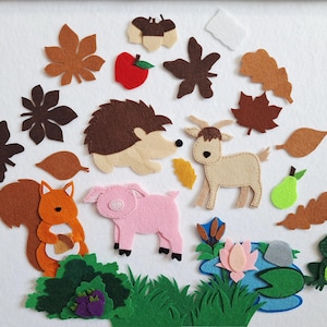 May include: A selection of felt animal and nature figures, including a squirrel, pig, goat, hedgehog, and frog. The scene also features leaves, acorns, an apple, and a pear, all in shades of brown, orange, green, and pink. A pond with a lily pad is also present.