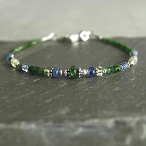 May include: A delicate bracelet featuring a mix of green, blue, and silver beads. The bracelet has a silver clasp and is strung on a flexible wire. The beads include small green stones, blue beads, and silver accents, creating a colorful accessory.