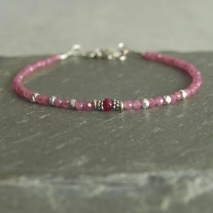 May include: A delicate pink gemstone bracelet with a silver clasp. The bracelet features small, round pink beads interspersed with silver-colored accents and a central cluster of darker pink beads. The bracelet is displayed on a gray surface.