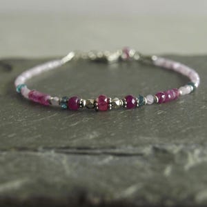 May include: Delicate beaded bracelet featuring a mix of pink, magenta, and silver beads. The bracelet includes small, faceted ruby beads, interspersed with silver-toned metal beads and small blue-green beads. The bracelet is strung on a thin, light pink strand.