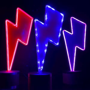 Może przedstawiać: Trzy neonowe lampy w kształcie błyskawicy w kolorze czerwonym, niebieskim i fioletowym. Każda błyskawica jest zamontowana na małej, ciemnej, cylindrycznej podstawie. Tło jest ciemne i jednolite, co podkreśla żywą poświatę świateł.