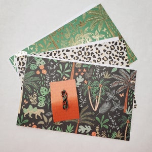 May include: A stack of four patterned wrapping papers with a green, gold, and white leopard print, a green and gold tropical print, and a black and green tropical print with a small orange tag with a green and black string.
