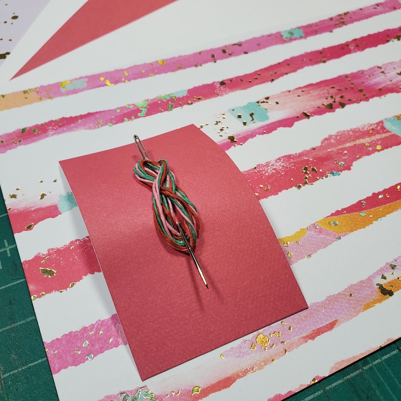 May include: A red card with a needle threaded with green, pink, and red embroidery floss. The card is on top of a white background with pink and gold stripes.