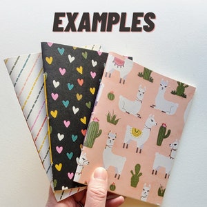 May include: Three small notebooks with different patterned covers. The covers are black with white hearts, pink with white llamas, and white with yellow stripes. The notebooks are stacked on top of each other and the word "EXAMPLES" is written in black letters on a white background above the notebooks.