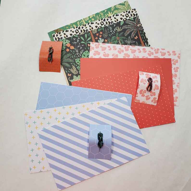 May include: A set of patterned scrapbook paper in various colors, including red, blue, green, and white. The paper features different patterns, including polka dots, stripes, and floral designs. There are also small decorative pieces, such as a black and green wire twist, on the paper.