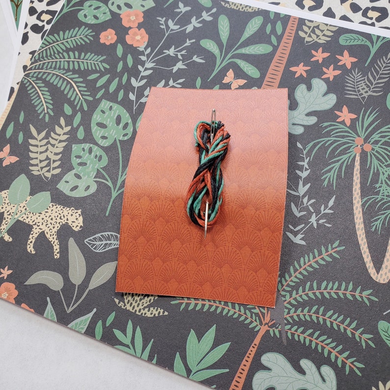 May include: A close-up of a needle threaded with black, green, and orange embroidery floss. The needle is resting on a piece of orange cardstock with a decorative pattern.