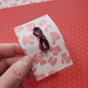 May include: A close-up of a needle threaded with black, orange, and purple embroidery floss on a white background with a pink leopard print.