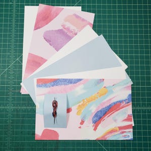 May include: A stack of colorful patterned paper sheets with a small red and black needle on a blue card. The paper sheets are arranged in a fan shape with the bottom sheet having a pink, blue, and yellow abstract design.