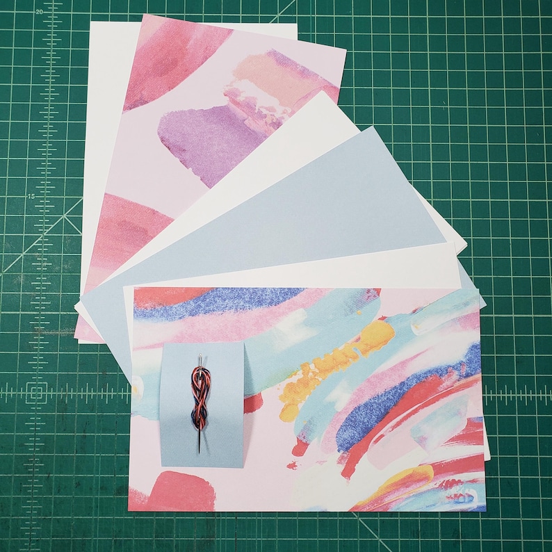 May include: A stack of colorful patterned paper sheets with a small red and black needle on a blue card. The paper sheets are arranged in a fan shape with the bottom sheet having a pink, blue, and yellow abstract design.
