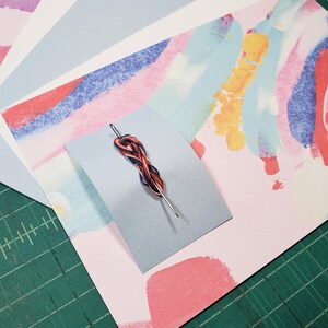 May include: A needle threaded with red, blue, and black embroidery floss on a light blue piece of paper with a pink and blue abstract watercolor background.