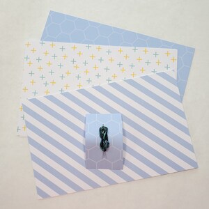 May include: Four sheets of patterned paper in shades of blue and white. The top sheet has a honeycomb pattern, the second sheet has a pattern of small yellow and green plus signs, the third sheet has a pattern of diagonal blue stripes, and the bottom sheet has a honeycomb pattern. A green thread is wrapped around a needle and placed on the third sheet.