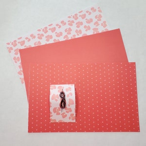 May include: Three sheets of red paper with different patterns. The top sheet has a white background with pink leopard print. The middle sheet is solid red. The bottom sheet has a white background with small pink polka dots. A small piece of brown string is on top of the sheets.