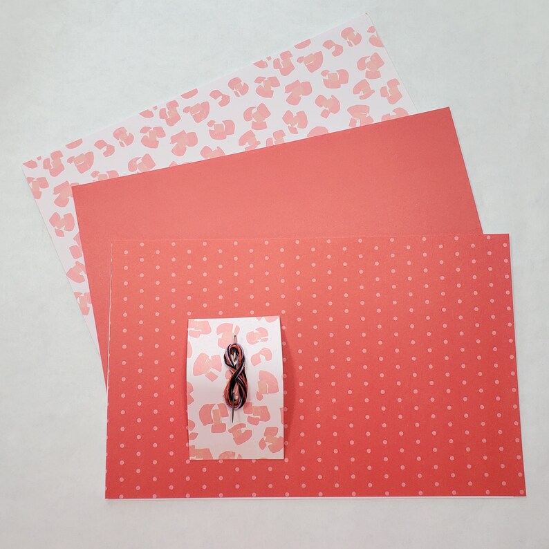 May include: Three sheets of red paper with different patterns. The top sheet has a white background with pink leopard print. The middle sheet is solid red. The bottom sheet has a white background with small pink polka dots. A small piece of brown string is on top of the sheets.