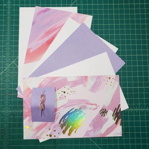 May include: A collection of pink and purple patterned paper sheets with gold accents. The paper is arranged in a fan shape with a small purple card with a pink and gold string tied in a knot on top.