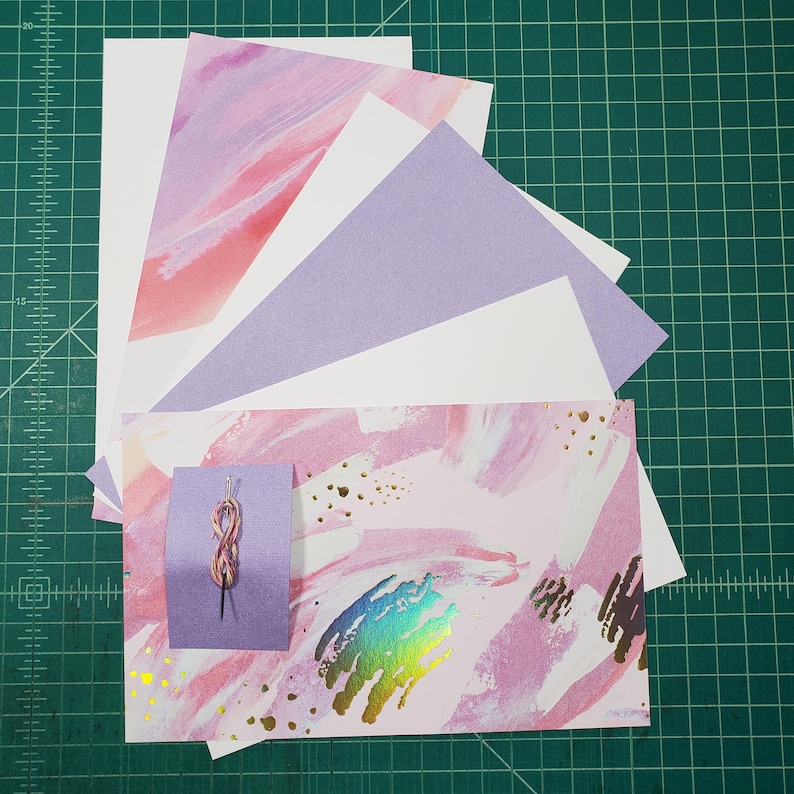 May include: A collection of pink and purple patterned paper sheets with gold accents. The paper is arranged in a fan shape with a small purple card with a pink and gold string tied in a knot on top.