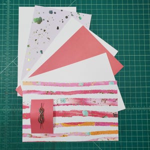 May include: A stack of colorful patterned paper with a pink and white striped paper on the bottom. A small red card with a brown string is on top of the striped paper.