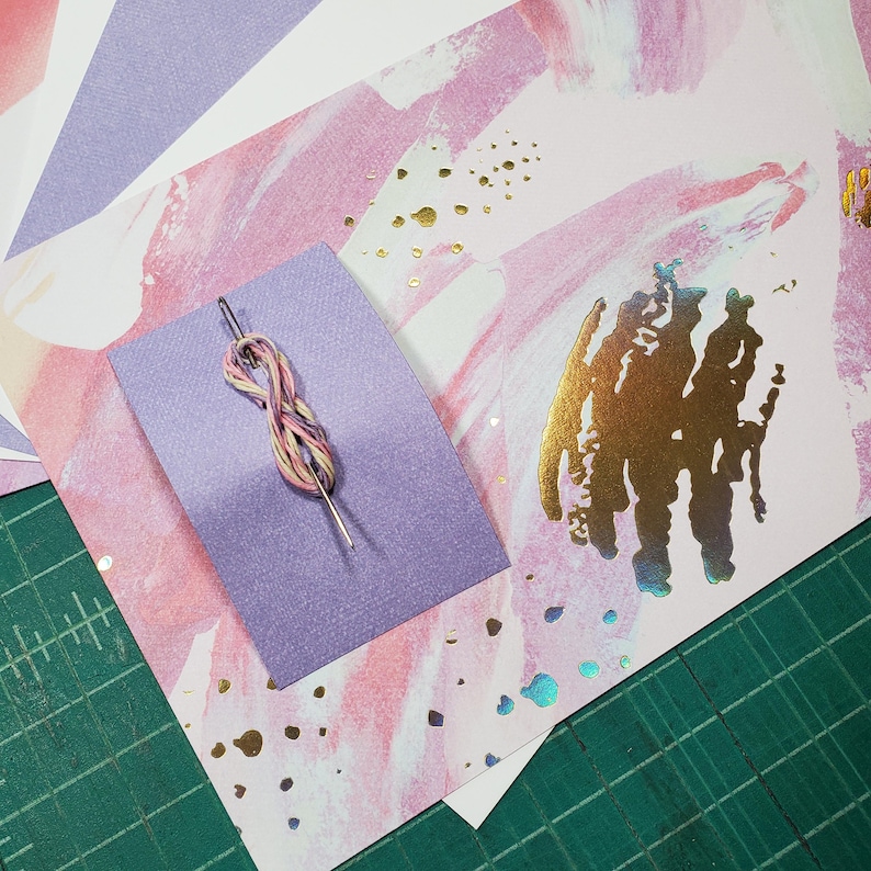 May include: A purple piece of fabric with a needle and thread on top. The fabric is on top of a white background with pink and gold abstract designs.