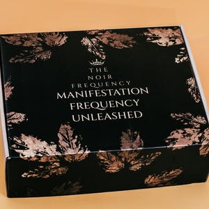 May include: A black box with a rose gold leaf pattern and the text "THE NOIR FREQUENCY MANIFESTATION FREQUENCY UNLEASHED" in white. The box is on a light orange surface.