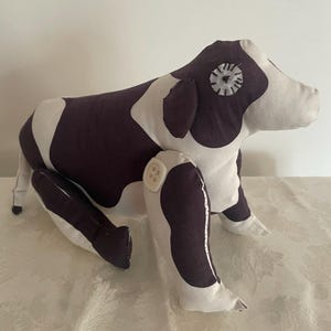 Puede incluir: Un juguete de peluche hecho a mano con forma de perro, en posición sentada. El juguete es blanco con manchas y detalles en morado oscuro. El perro tiene articulaciones de botón y un ojo de fieltro. El juguete es de tela suave.