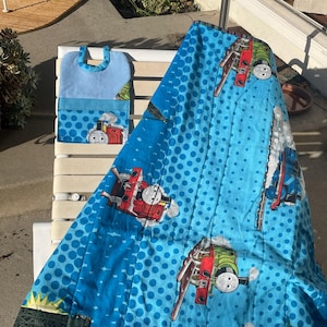 May include: A blue and white Thomas the Tank Engine themed quilt and matching tote bag. The quilt features a repeating pattern of the train on a blue background with white polka dots. The tote bag is light blue with a Thomas the Tank Engine graphic.
