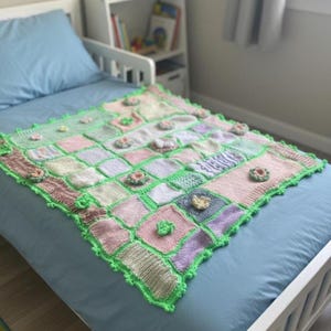 May include: A handmade, square patchwork blanket in pastel colors of pink, green, and lavender. The blanket features a green border and is adorned with small, three-dimensional flower appliqués. The blanket is displayed on a blue bed.