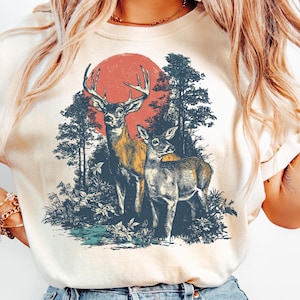 May include: Cream-colored t-shirt featuring a vintage-style illustration of two deer in a forest setting. A large red sun is in the background. The deer are in shades of brown and gray, with detailed antlers and fur.