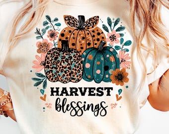 May include: Cream-colored t-shirt featuring a fall-themed graphic. The design includes three pumpkins in shades of brown, teal, and leopard print, surrounded by floral accents. The words "HARVEST blessings" are printed in black script.