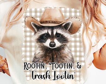May include: Cream-coloured t-shirt featuring a raccoon wearing a cowboy hat. The raccoon is set against a tan and white checkered background. The text "Rootin', Tootin', & Trash Lootin'" is printed below the raccoon.