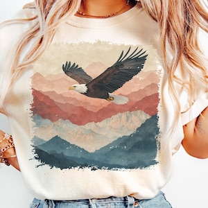 Op de afbeelding: Cr&egrave;mekleurige sweatshirt met een zwevende Amerikaanse zeearend met uitgestrekte vleugels tegen een achtergrond van gelaagde bergen in tinten rood, blauw en beige. De adelaar heeft bruine veren, een witte kop en een gele snavel.