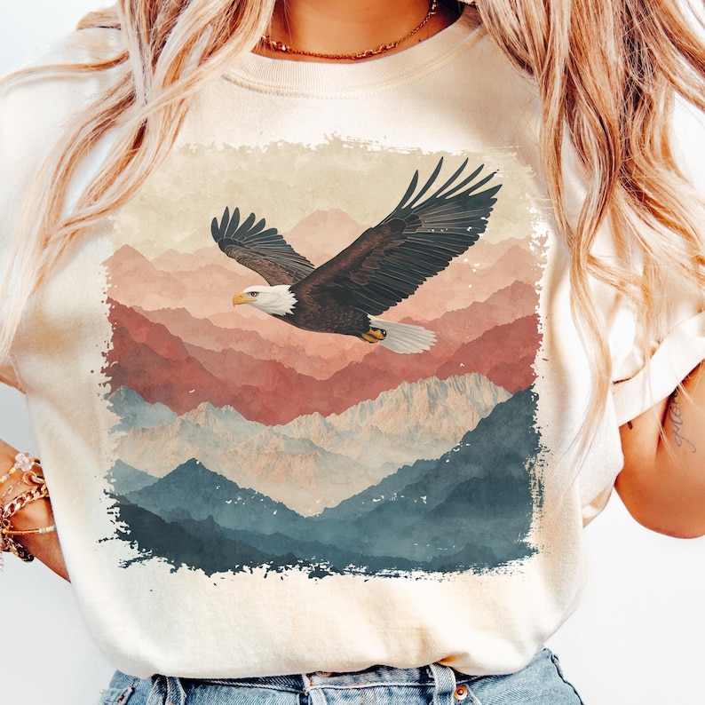 Op de afbeelding: Cr&egrave;mekleurige sweatshirt met een zwevende Amerikaanse zeearend met uitgestrekte vleugels tegen een achtergrond van gelaagde bergen in tinten rood, blauw en beige. De adelaar heeft bruine veren, een witte kop en een gele snavel.