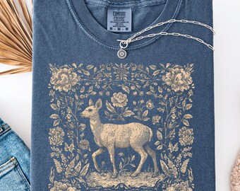 Op de afbeelding: Een marineblauw t-shirt met een hertenafbeelding omringd door bloemmotieven. Het shirt heeft een ronde hals en korte mouwen. Een zilveren ketting met een bloemenhanger is over het shirt gedrapeerd. Het label luidt "COMFORT COLORS".