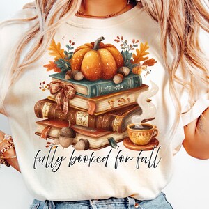 May include: Cream-colored t-shirt featuring a fall-themed graphic. A stack of books is topped with a pumpkin, acorns, and fall foliage. A cup of coffee sits beside the books. The text "fully booked for fall" is written in a script font.