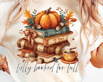 May include: Cream-colored t-shirt featuring a fall-themed graphic. A stack of books is topped with a pumpkin, acorns, and fall foliage. A cup of coffee sits beside the books. The text "fully booked for fall" is written in a script font.