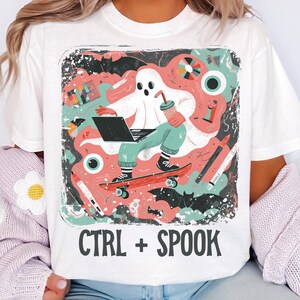 May include: White t-shirt with a colourful graphic of a ghost on a skateboard using a laptop and drinking a beverage. The design includes bats, eyes, and the text "CTRL + SPOOK".