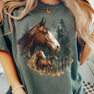 May include: A green t-shirt with a detailed illustration of three horses in a forest. The design features a large horse head and two smaller horses, set against a backdrop of trees. The horses are brown with white markings.