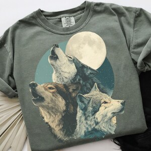 May include: A sage green sweatshirt featuring a graphic of three wolves howling at a full moon. The wolves are depicted in shades of brown, gray, and white, set against a teal and navy blue background. The moon is a bright cream color.