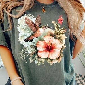 May include: Dark green t-shirt featuring a vibrant illustration of a hummingbird in flight, set against a backdrop of tropical flowers and foliage. The design includes a large, peach-colored hibiscus flower.