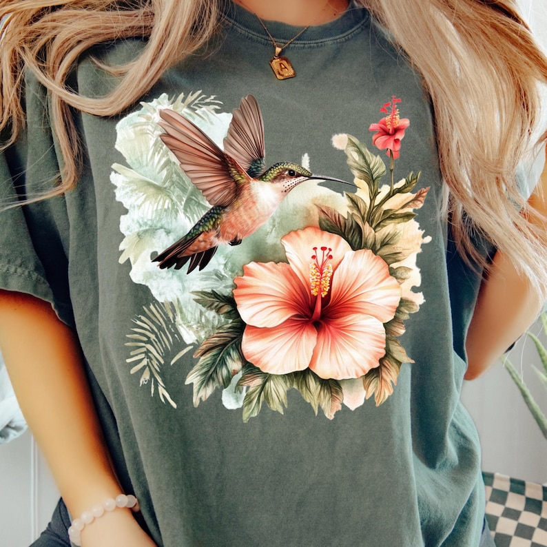 May include: Dark green t-shirt featuring a vibrant illustration of a hummingbird in flight, set against a backdrop of tropical flowers and foliage. The design includes a large, peach-colored hibiscus flower.