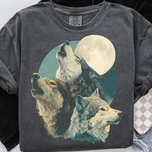 May include: Dark gray sweatshirt featuring a graphic of three wolves howling at a full moon. The wolves are depicted in shades of brown, gray, and white, set against a teal and navy blue background. The sweatshirt has a crew neck and long sleeves.