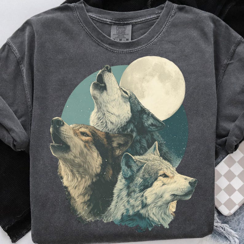 May include: Dark gray sweatshirt featuring a graphic of three wolves howling at a full moon. The wolves are depicted in shades of brown, gray, and white, set against a teal and navy blue background. The sweatshirt has a crew neck and long sleeves.