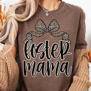 May include: A brown t-shirt featuring the words "Foster Mama" in a cursive font, with a leopard print bow design above the text. The shirt has short sleeves and a crew neck.