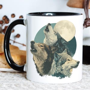 May include: White ceramic mug with a black handle and rim. The mug features a detailed illustration of three wolves howling at a full moon. The background includes a teal circle with stars.