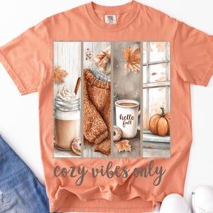 Pode incluir: Camiseta cor de p&ecirc;ssego com um gr&aacute;fico tem&aacute;tico de outono. O gr&aacute;fico inclui um caf&eacute; com leite, um su&eacute;ter, uma caneca de caf&eacute; com "hello fall" e uma ab&oacute;bora. O texto "cozy vibes only" est&aacute; impresso abaixo.